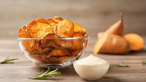 Delicious sweet potato chips in bowl, rosemary and sauce on table