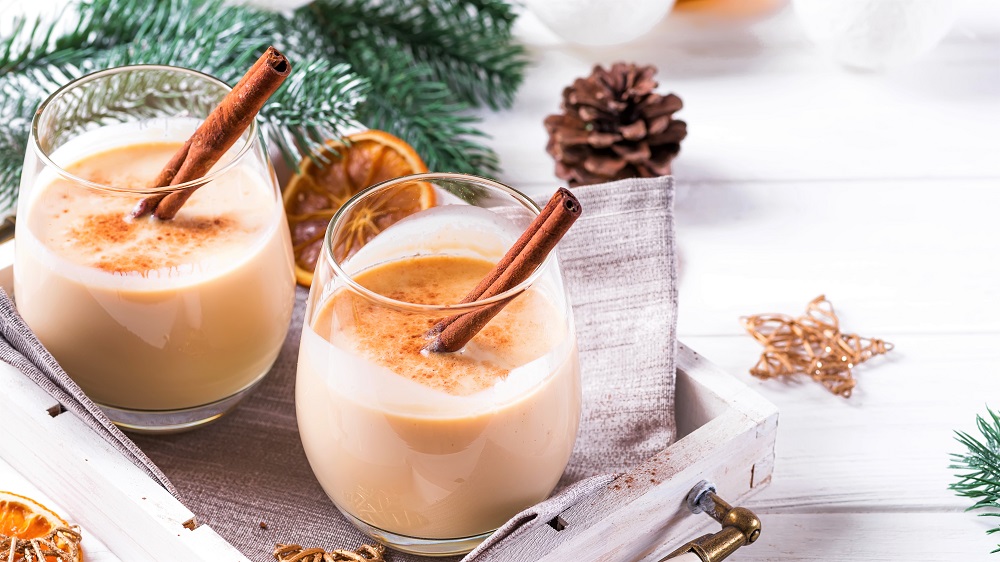 Eggnog in glasses with cinamon in tray, dry orange,fir branches and pine cone on white wooden table