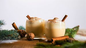 selective focus of eggnog cocktail with whipped cream near cinnamon sticks and spruce branches on wooden table isolated on grey