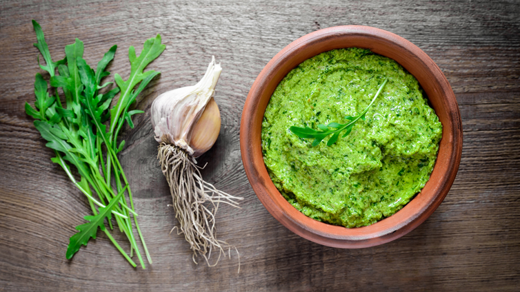 Rucola Pesto in kleinem Tontopf neben Bund Rucola und Knoblauch