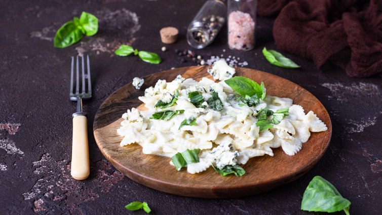 Farfalle in Gorgonzola-Soße sind mit Basilikum-Blättern auf einem Holzteller angerichtet.