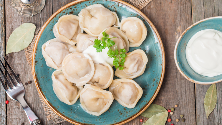 Pelmeni sind mit Schmand und Petersilie auf einem blauen Teller angerichtet.